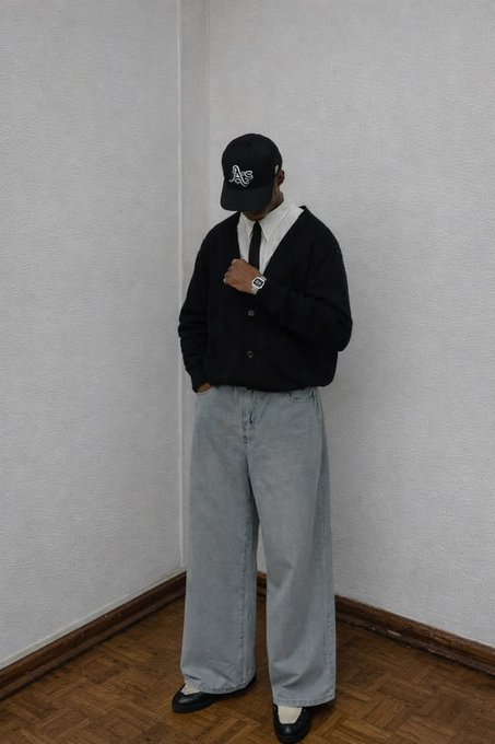 A person poses in a corner wearing a black cap with 'AF' lettering, a white collared shirt with tie, a black cardigan, baggy light blue jeans, and black and white shoes.