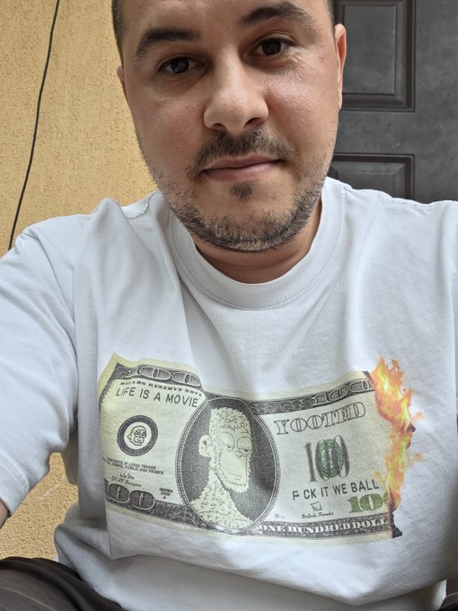 A man wearing a white t-shirt featuring a burned $100 bill design with Rick Sanchez's face and the text "LIFE IS A MOVIE" and "F CK IT WE BALL" takes a close-up selfie, matching the post's playful crypto culture reference to the y00ts and DeGods NFT communities.