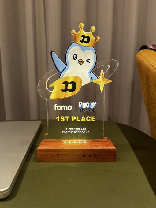 A 1st place acrylic trophy featuring a crowned penguin mascot with a winking eye, displayed on a wooden base. The award reads 'fomo | Pudgy Penguins 1ST PLACE - A TRADING APP FOR THE REST OF US' and is inscribed with 'KRIM 2026' and 'fomo' on the base.