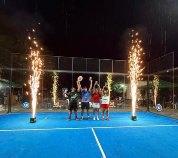 The image features a group of four people standing together on a tennis court, celebrating their victory with raised arms. They are surrounded by several large fireworks that have been set off for them, creating an exciting atmosphere. The background includes a blue tennis court and some chairs scattered around the area. The post accompanying this image is titled "Los campeones del Cake Latam Cup ellos se llevaron el premio en @monero tres días de puro padel, adopción cripto, educación y economía circular Gracias @cakewallet @TrocadorApp Por este mágico evento ccripto sport!".