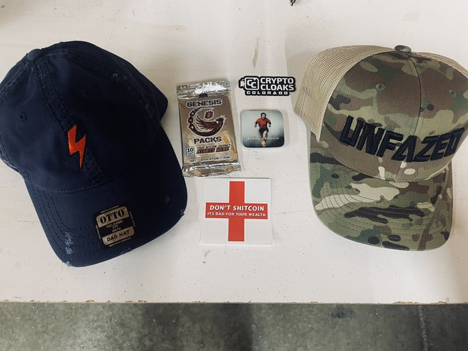 A flat lay of merchandise including a navy 'OTTO DAD HAT', a camouflage 'UNFAZED' hat, a pack of Genesis 'Sovereign' rolling papers, Crypto Cloaks and 'DON'T SHITCOIN' stickers, and a runner button pin.