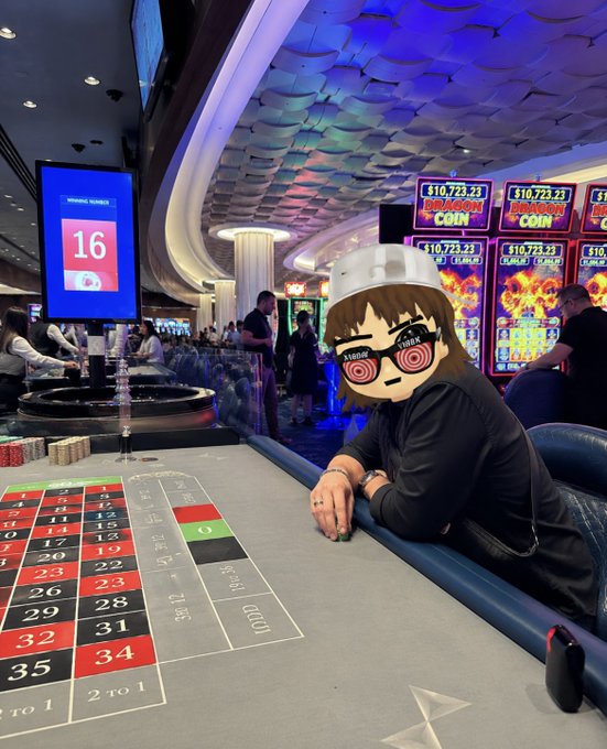 A person wearing a black shirt and a metallic headpiece sitting at a roulette table in a casino, with their face obscured by a cartoon emoji wearing sunglasses with "SWAG" and "CREW" text; slot machines and other patrons are visible in the background.