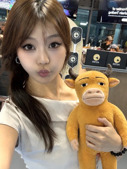 A young woman with long brown hair wearing a white top poses with a pouty expression while holding an orange plush toy with horns in what appears to be an ESTER arcade or gaming venue, with visible signage reading "ESTER" and "The King's Landing" in the background.