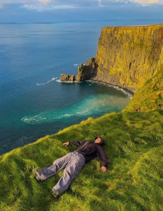 A person lying on a grassy cliff edge overlooking turquoise ocean waters and dramatic coastal cliffs.