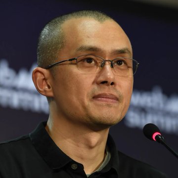 A close-up photo of a man with short hair and glasses speaking into a microphone, wearing a black shirt, with a blurred dark background.