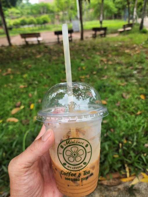 The image features a person holding a cup of coffee with a straw, which has the logo "Mai coffee Thuốc tôi hút : Sài gòn bạc Kèo tôi quan tâm @quipnetwork Ae cùng quan điểm cmt bàn luận vui vẻ!" The person is holding the cup in their hand, and there are benches visible in the background.