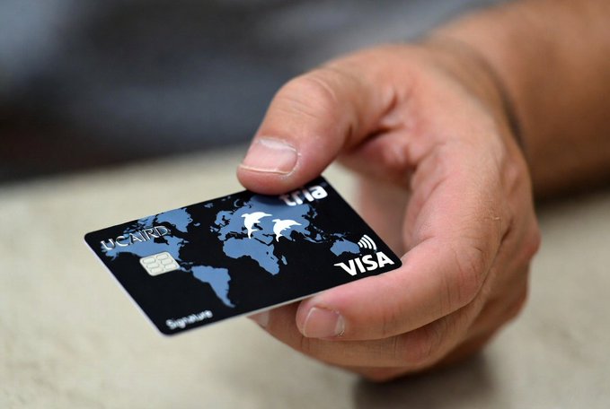 The image shows someone's hand holding up a black Visa credit card with a white picture of a dog, as well as some writing around the bottom right corner of the card. The hand appears to be in close proximity to another person who seems to be looking at their phone.