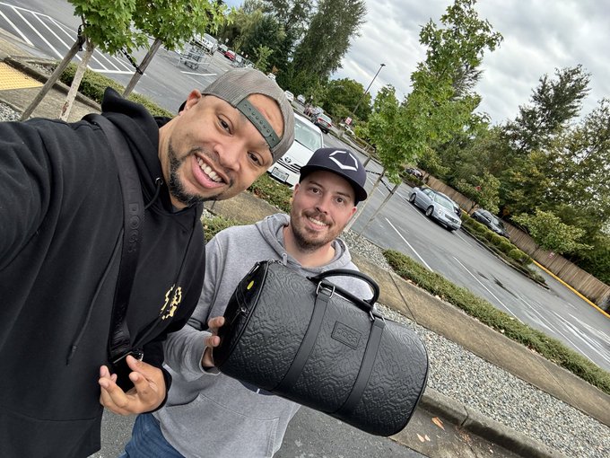 Two smiling men pose for a selfie in a parking lot, with one wearing a black hoodie and the other a gray hoodie and baseball cap while holding a black textured duffel bag.