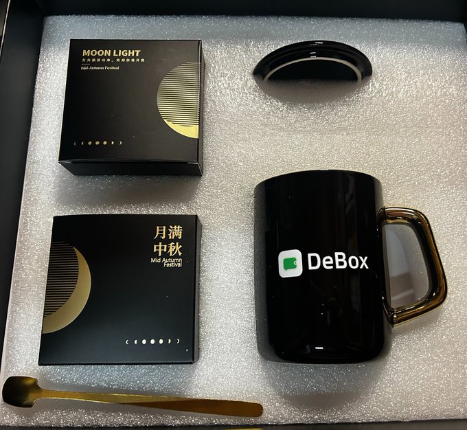 A 'MOON LIGHT' Mid-Autumn Festival gift set in a black presentation box with moon phase designs, containing a black DeBox-branded mug with gold handle and a gold spoon.