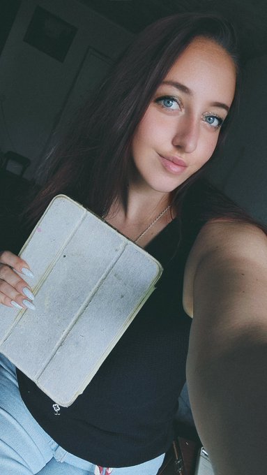 A young woman with long dark hair holds up a tablet or notebook while taking a selfie in a dimly lit room.
