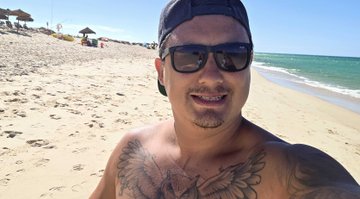 A shirtless man wearing sunglasses and a backwards cap takes a selfie on a sunny beach, with visible chest tattoos. The casual beach setting contrasts with his described role as a Web3 founder and property landlord, suggesting a moment of leisure away from his entrepreneurial and real estate work.