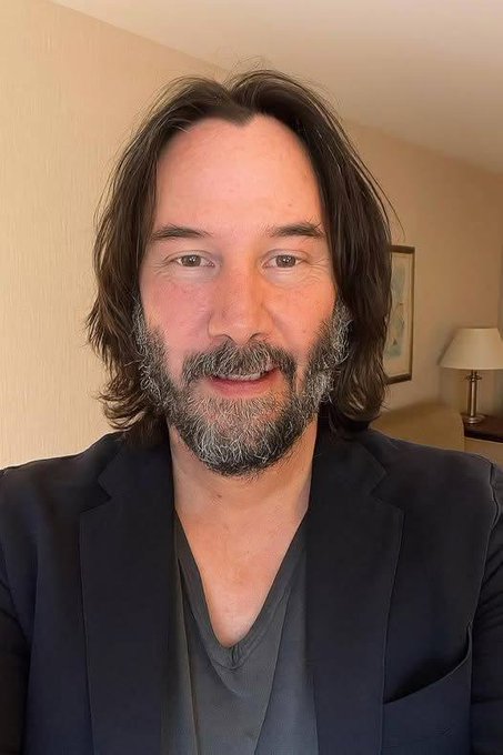 A man with shoulder-length dark hair and a salt-and-pepper beard wears a black blazer over a dark shirt, photographed indoors.
