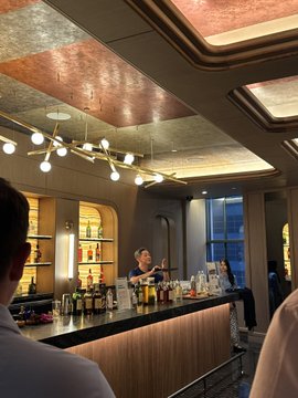 The interior of an upscale bar with a bartender standing behind a counter stocked with bottles, featuring an ornate ceiling with multiple modern globe pendant lights.