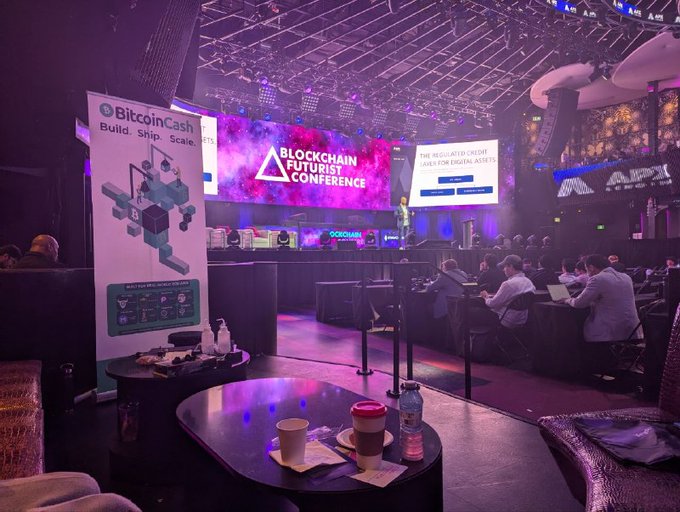 A view of a 'BLOCKCHAIN FUTURIST CONFERENCE' with a BitcoinCash banner ('Build. Ship. Scale.'), purple lighting, a presentation screen, and audience members seated at tables.