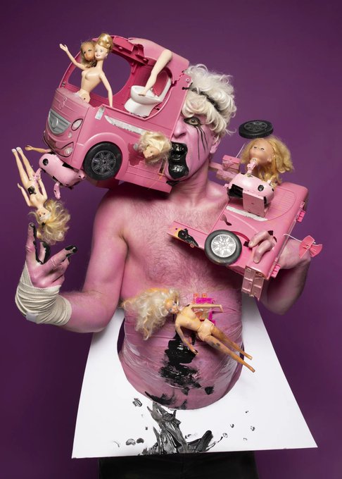 A surreal artistic photograph features a shirtless man with pink-painted skin wearing blonde wigs and surrounded by broken Barbie dolls and pink toy cars against a purple background.