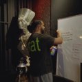 A small dark image showing a person wearing a green 'Eat' shirt near what appears to be a whiteboard with writing.