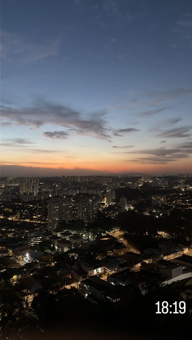 A scenic dusk view of a sprawling city skyline with lights beginning to twinkle in the buildings and streets, under a sky transitioning from orange near the horizon to deep blue above, with a timestamp of '18:19' in the corner.