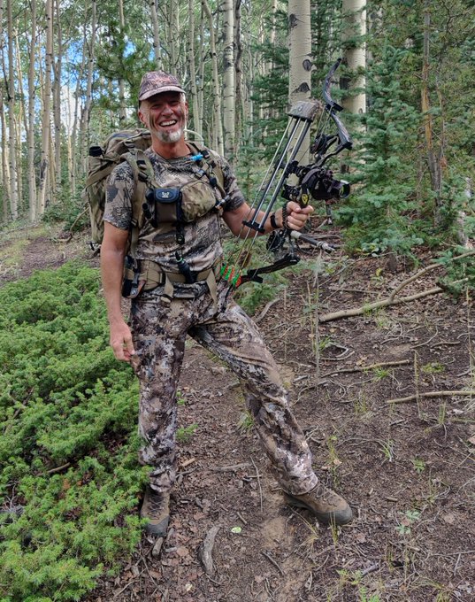 A smiling man in full camouflage hunting gear standing in a forest, holding a compound bow and wearing a backpack.
