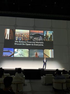 A man presents on stage at a conference with a large screen displaying 'We Believe Financial Markets Should Be Open to Everyone' alongside various images.