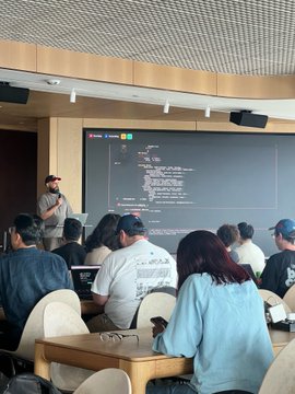 The image shows a group of people sitting around a table, with one person standing at the front giving a presentation or lecture. The attendees are using laptops and cell phones while listening attentively. In the background, there's a large screen displaying information related to Capacitr running MiroShark simulations during a @base hackathon event.