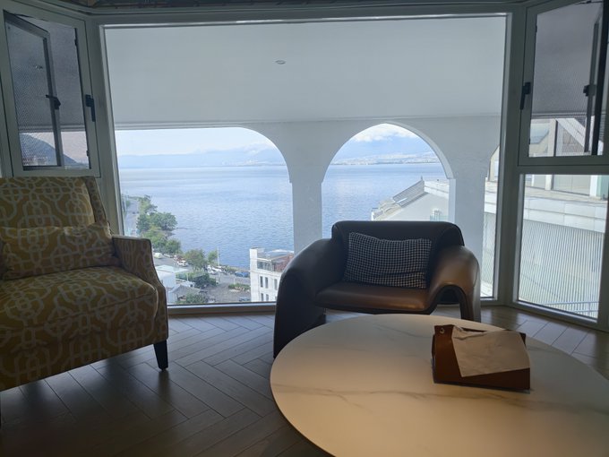 A bright, seaside lounge area with arched windows overlooking the ocean features a patterned yellow armchair, a black chair with a checkered pillow, and a round table with a small bag. The serene coastal view contrasts ironically with the post's chaotic tone of chasing multiple meme coins ($牛来, $cashcat, $PONS, $Chonketha) — a calm setting masking the frenzy of speculative crypto trading.