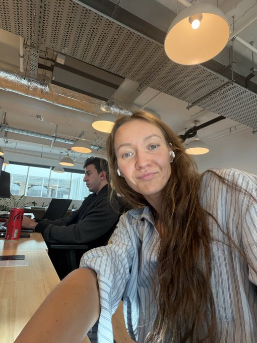 A woman with long brown hair, wearing earbuds and a striped shirt, smiles at the camera while taking a selfie in an open office space with exposed ceilings and pendant lighting. In the background, a man in a dark shirt works intently on his laptop at a desk with a red Coca-Cola can visible, matching the post's reference to a busy colleague at the Coinbase UK office.