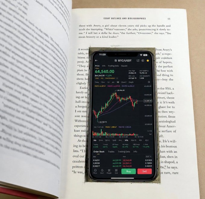 A smartphone displaying a BTC/USDT trading chart with a price of $64,540.00 rests on an open book titled 'ESSAY OUTLINES AND BIBLIOGRAPHIES'.