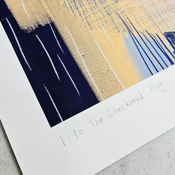 A close-up of an art print's bottom corner showing abstract blue and beige vertical brushstrokes with handwritten text reading '1/10 The Checkered Hug'.