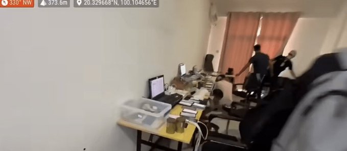 A small, cluttered office or workspace with desks, computers, and people visible through what looks like a security camera or first-person view, with overlay text showing coordinates '330° NW', '373.6m', and '20.329668°N, 100.404656°E'.