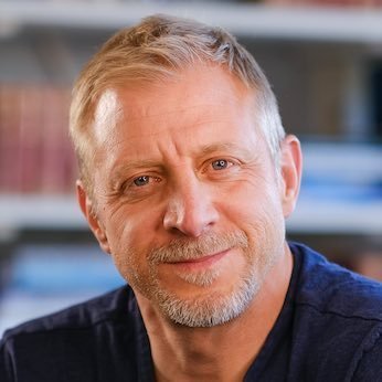 A headshot portrait of a middle-aged man with short graying hair and a goatee, wearing a dark shirt, pictured against a softly blurred bookshelf background. The accompanying quote from James Lavish expresses his conviction that Bitcoin's current price below $100K is a rare opportunity that future generations will view as legendary.