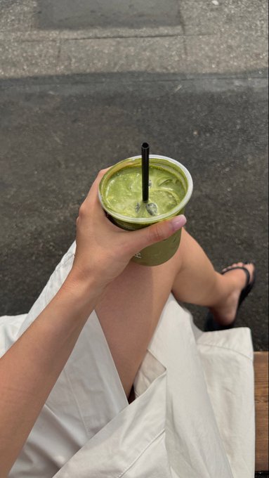 A hand holds a frothy iced matcha latte with a black straw, the bright green drink contrasting against the white outfit and gray pavement below, capturing a calm slow Sunday moment.