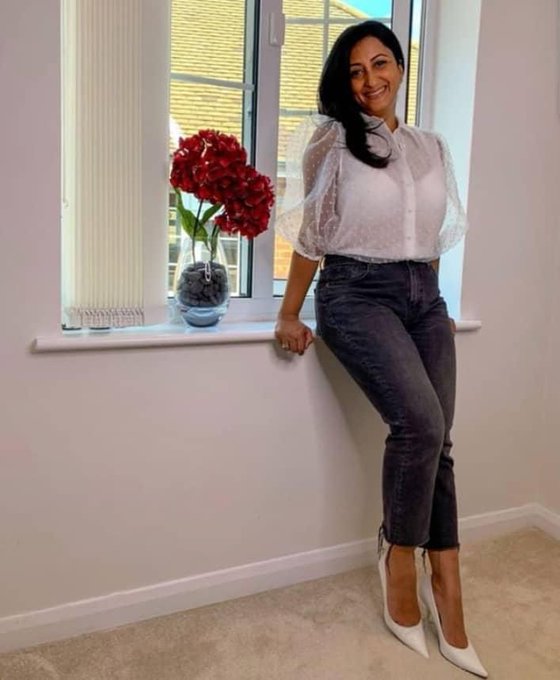 The image features a woman standing next to a window with her legs crossed. She is wearing a white blouse and black pants, and she is smiling at the camera. In front of her are two potted plants - one red and one white. The background includes a wall painted in white, which contrasts nicely with the vibrant colors of the flowers on display. There is also a vase placed near the window, adding to the overall aesthetic of the scene.