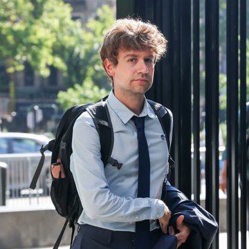 A man with messy hair wearing a light blue dress shirt, a dark blue tie, and a black backpack stands on a city street.