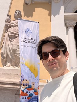 A man wearing sunglasses and a white t-shirt standing in front of a stone building with a banner and a statue.
