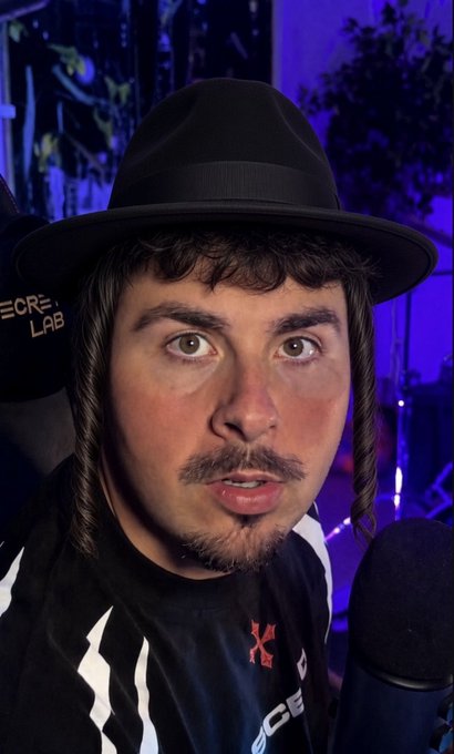 A young man with long sidelocks and a black fedora speaks into a microphone, wearing a black shirt with white stripes and a red "K" logo. Behind him, "SECRET LAB" text is partially visible against a blue-lit background, fitting the casual "gm" greeting to crypto Twitter followers.