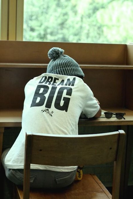 A person wearing a gray beanie sits with their head down at a wooden desk, wearing a white t-shirt with 'DREAM BIG' printed on the back, with sunglasses on the desk nearby.