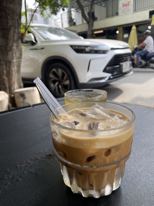 A glass of iced coffee on a black table in the foreground, with a white SUV parked on a Vietnamese street visible in the background.