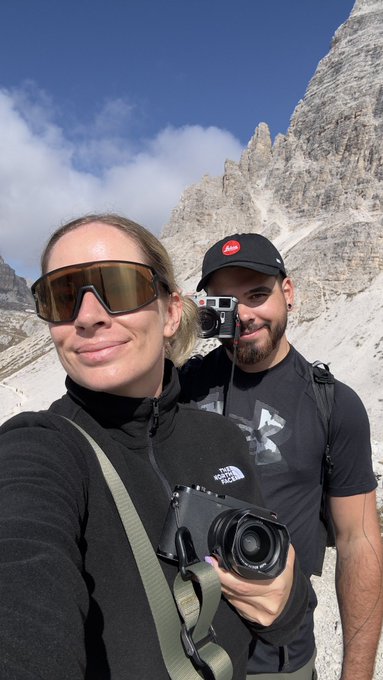 A couple takes a selfie in a mountainous landscape under a blue sky with clouds; the woman in the foreground wears sunglasses and a black North Face fleece holding a camera, while the man behind her wears a black cap and a t-shirt with a GoPro mounted on his chest strap.