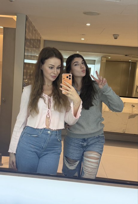 Two young women take a mirror selfie in what appears to be a bathroom or hallway; the one on the right makes a playful kiss face and peace sign.