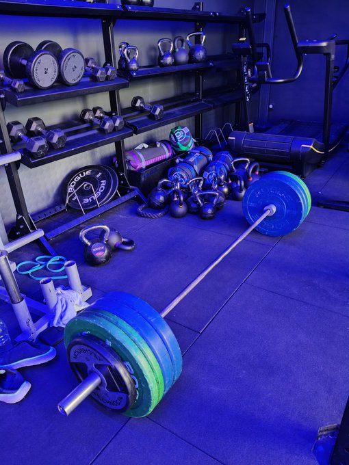 A home gym setup is bathed in blue lighting, featuring a loaded Rogue barbell with colorful bumper plates ready for deadlifts, alongside organized racks of dumbbells, kettlebells, weight plates, and a battle rope. The scene perfectly complements the post's morning greeting and question about deadlifting, showcasing the essential equipment needed for the lift.