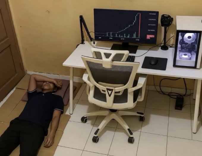 A man lies on a pillow on the floor beneath a white desk that holds a computer monitor displaying a rising stock chart, a PC tower with purple lighting, and a white ergonomic office chair.