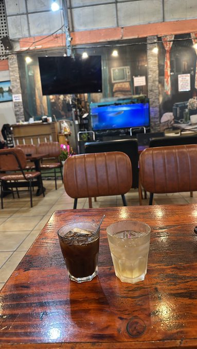 Two iced drinks, one dark coffee and one lighter beverage, on a wooden table inside a café with leather chairs and TV screens in the background.