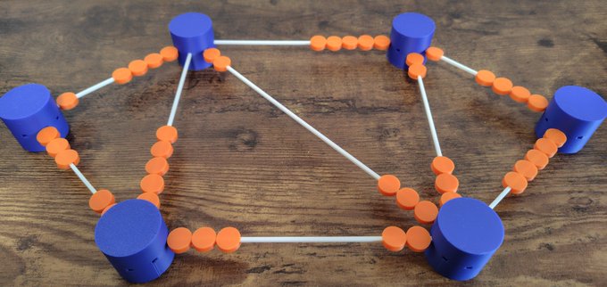 The image features a wooden table with a unique Lego structure composed of blue and orange circles, resembling a network or grid-like pattern. The Lego pieces are connected by white lines, creating an intricate design that resembles a lightning network model. The text in the image reads "At Tokyo Bitcoin Base, we are displaying the Lightning Network Model" and "Focus on what is in the image and how it relates to the post".