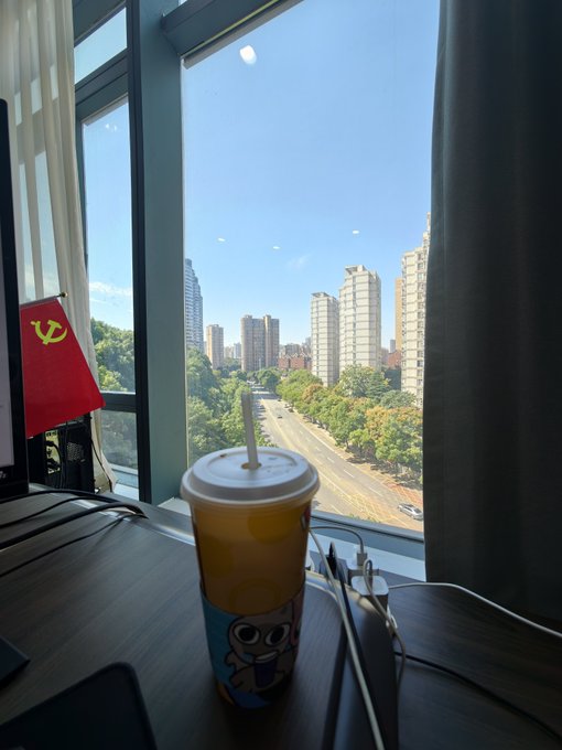 An iced drink sits on a wooden desk by a window that frames a sunny city skyline of high-rise buildings, capturing the early morning reset before the start of TOKEN2049. A small red flag with a yellow hammer and sickle emblem rests nearby, adding a subtle pop of color to the scene.