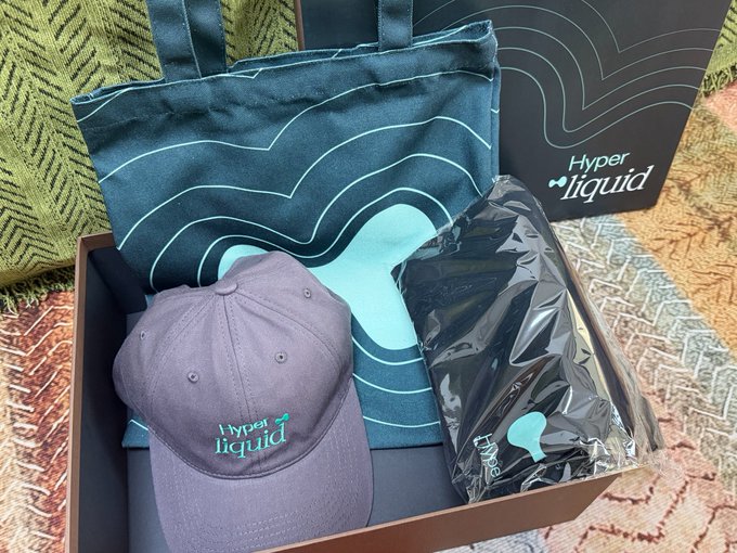 A physical merchandise box set from 'Hyper liquid' containing a dark blue patterned tote bag, a grey cap with the 'Hyper liquid' logo, and an unidentified item wrapped in shiny black plastic.