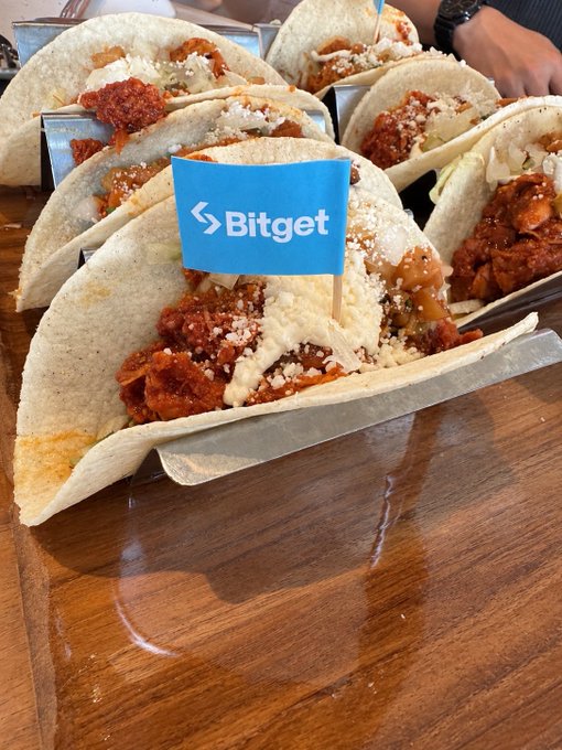 Several soft tacos filled with fried chicken, coleslaw, and sauce, displayed in metal holders on a wooden table with a small blue 'Bitget' flag inserted in the center.