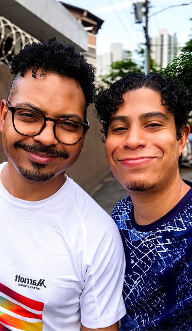 Two smiling young men pose for a selfie on a street; one wears glasses and a white t-shirt with a colorful 'Marathon' logo, while the other wears a blue patterned shirt.