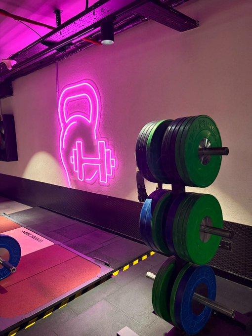 A gym workout area featuring a pink neon sign of a kettlebell and dumbbell on the wall, alongside a rack of green, blue, and black weight plates and a lifting platform.