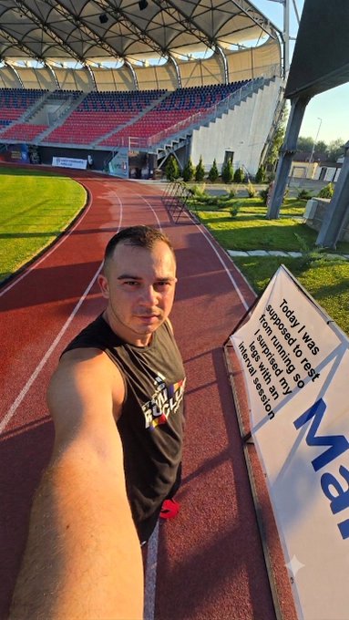 A man takes a selfie at a running track stadium, with a sign reading 'Today I was supposed to rest I am running so I targeted my interval with this session' and 'Max' visible.