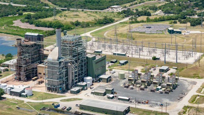 An aerial view of an industrial facility or power plant with smokestacks, storage tanks, and an electrical substation surrounded by green fields.